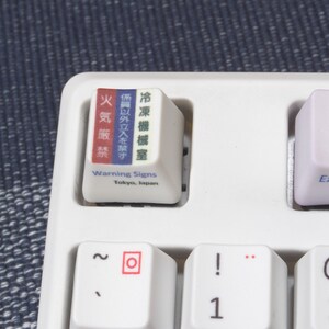 May include: A white keyboard with a keycap that has Japanese text and the words "Warning Signs" and "Tokyo, Japan". The keycap is red, white, and blue. The keyboard also has keys for the numbers 1 and 2, the letters "F1", "@", and "-", and the word "Explorer".