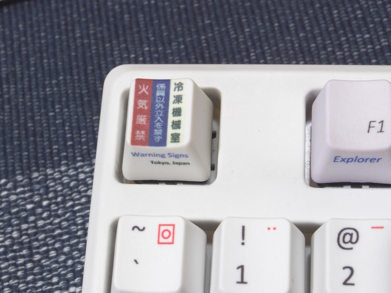 May include: A white keyboard with a keycap that has Japanese text and the words "Warning Signs" and "Tokyo, Japan". The keycap is red, white, and blue. The keyboard also has keys for the numbers 1 and 2, the letters "F1", "@", and "-", and the word "Explorer".