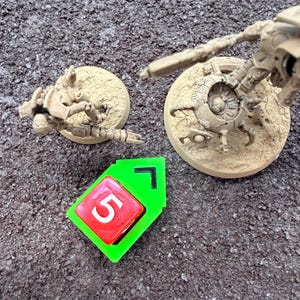 May include: An overhead shot shows a green and red game piece with the number 5, and two beige miniature figurines on circular bases. The figurines appear to be part of a tabletop game, set against a textured, dark background.