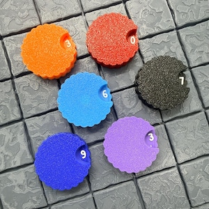 Pode incluir: Uma coleção de fichas de jogo circulares e coloridas com bordas onduladas. Cada ficha tem uma cor diferente: laranja, vermelho, azul, preto e roxo. Os números brancos são impressos em cada ficha: 0, 3, 6, 7 e 9. As fichas estão em uma superfície cinza texturizada.