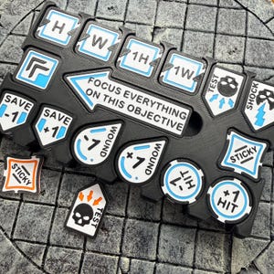 May include: A black plastic game board featuring a large arrow with the text FOCUS EVERYTHING ON THIS OBJECTIVE. The board contains various white and blue game tokens with labels including SAVE, WOUND, HIT, STICKY, TEST, SHOCK, H, W, 1H, and 1W.