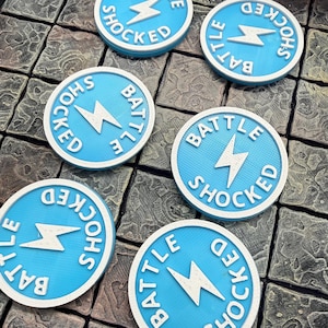 May include: Six circular light blue tokens featuring a white lightning bolt icon in the centre and the text BATTLE SHOCKED embossed in white lettering around the perimeter. Each token has a textured surface and a raised white border edge.
