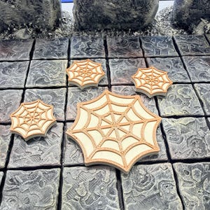 May include: Four beige and brown spiderweb-shaped game pieces. The pieces are arranged on a gray, stone-like surface. The spiderwebs have a detailed design with a light beige center and a brown border.