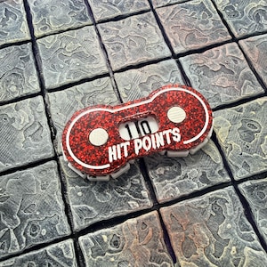 May include: A red glittery hit point counter with the number 10 displayed. The text "HIT POINTS" is printed in white. The counter is white-edged and sits on a gray, textured surface.