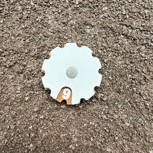 May include: A white, cog-shaped magnet with a silver centre and an orange accent displaying the number 9. The magnet has a textured surface and is set against a brown, speckled background.