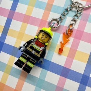 Könnte beinhalten: Ein Feuerwehrmann-Schlüsselanhänger mit einer Lego-ähnlichen Figur in schwarzer Uniform mit gelben und roten Akzenten, einem gelben Helm und einem kleinen orangefarbenen Flammenanhänger. Der Schlüsselanhänger ist an einer silbernen Kette und einem Ring befestigt.