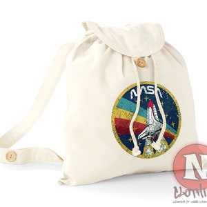 May include: A white canvas drawstring backpack with a vintage NASA logo featuring a space shuttle and the text "NASA" in blue. The logo has a red, yellow, blue, and green color scheme.