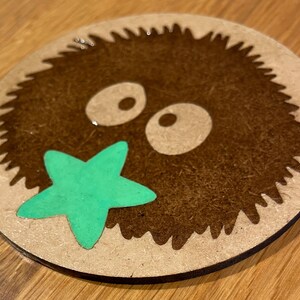 May include: A brown and white wooden coaster featuring a cartoon image of a furry creature with large eyes and a green starfish in its mouth.