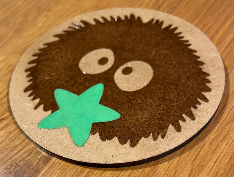 May include: A brown and white wooden coaster featuring a cartoon image of a furry creature with large eyes and a green starfish in its mouth.