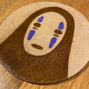 May include: A round, brown wooden coaster with a white background featuring a stylised illustration of No Face from the animated film Spirited Away. The illustration is in brown and purple.