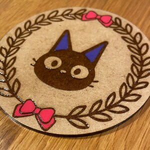 May include: A round wooden coaster featuring a brown cat illustration wearing a blue bow. The cat is surrounded by a brown wreath with pink bows on either side.