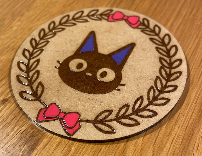 May include: A round wooden coaster featuring a brown cat illustration wearing a blue bow. The cat is surrounded by a brown wreath with pink bows on either side.