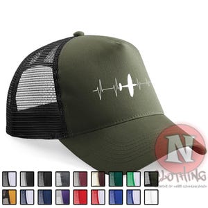 Peut inclure: Une casquette de type trucker vert olive avec un motif blanc représentant la silhouette d'un avion intégrée dans une ligne de rythme cardiaque EKG. La casquette possède un panneau avant structuré et un filet noir à l'arrière.