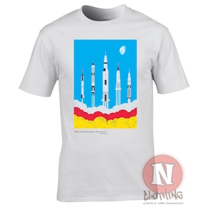 Puede incluir: Camiseta blanca con un gráfico de cinco cohetes lanzándose a un cielo azul con nubes blancas. La cita "We choose to go to the moon..." está impresa debajo del gráfico.
