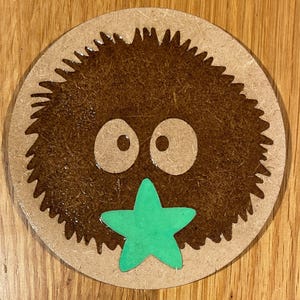 May include: A brown fuzzy round creature with two large eyes and a green star-shaped mouth. The creature is on a round wooden surface.