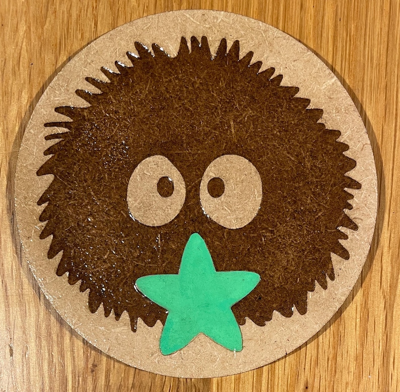 May include: A brown fuzzy round creature with two large eyes and a green star-shaped mouth. The creature is on a round wooden surface.
