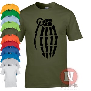 Puede incluir: Camiseta verde oliva con un diseño negro de granada de mano esquelética. Se muestran camisetas adicionales en varios colores a la izquierda. La camiseta tiene cuello redondo y mangas cortas. El logotipo de la marca es visible a la derecha.