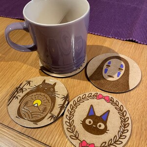 May include: Four round wooden coasters featuring different designs from the animated film 'Spirited Away'. The coasters depict No Face, Totoro, and the black cat from the film. The coasters are on a wooden table next to a purple mug.