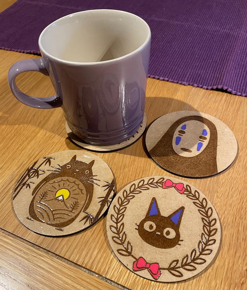 May include: Four round wooden coasters featuring different designs from the animated film 'Spirited Away'. The coasters depict No Face, Totoro, and the black cat from the film. The coasters are on a wooden table next to a purple mug.