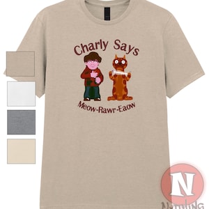 May include: A tan t-shirt featuring a cartoon illustration with the text "Charly Says" above a boy and a cat. The cat holds a comb, and the text "Meow-Rawr-Eaow" is below the figures. The shirt has a crew neck.