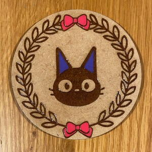 May include: A brown wooden coaster featuring a black cat silhouette in the centre. The cat has purple ears and large, round eyes. The cat is surrounded by a brown wreath with pink bows at the top and bottom.