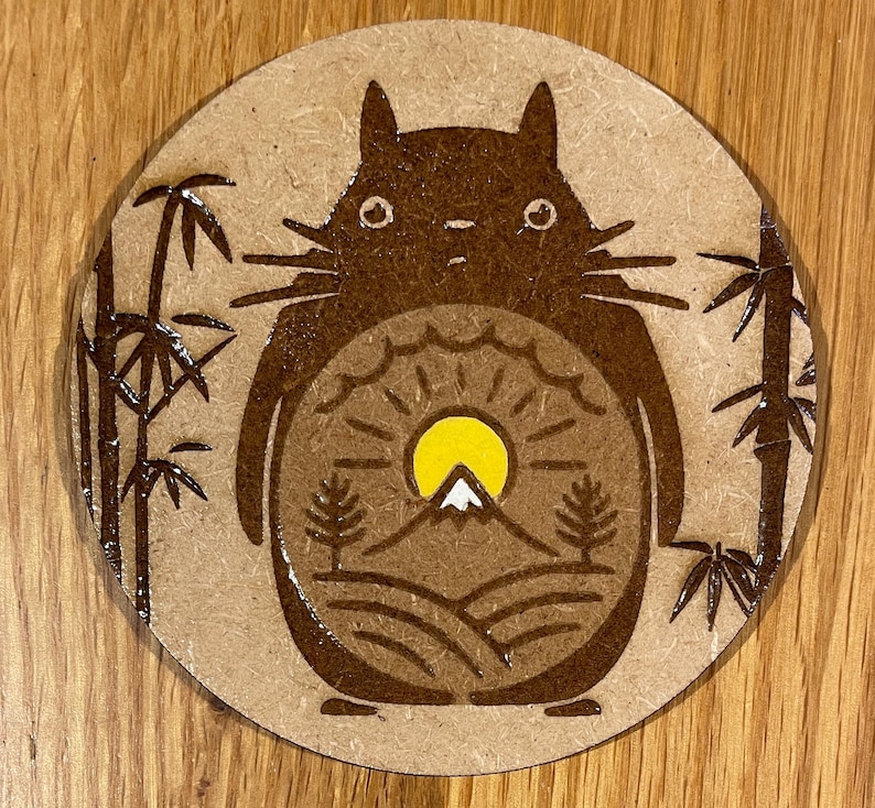 May include: A brown and white ceramic coaster featuring a cartoon character, a stylised mountain scene, and bamboo plants. The character has large, round eyes and a fluffy body.
