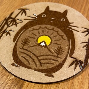 May include: A brown wooden coaster featuring a black and white illustration of a cartoon character with a mountain and sun inside. The character is surrounded by bamboo plants.
