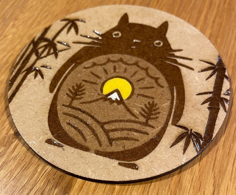 May include: A brown wooden coaster featuring a black and white illustration of a cartoon character with a mountain and sun inside. The character is surrounded by bamboo plants.