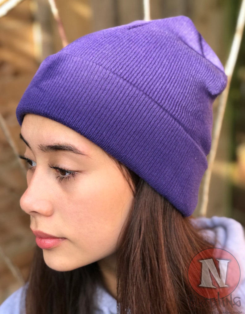 May include: A purple beanie hat made of knitted fabric. The hat is folded down at the brim and has a slouchy fit.