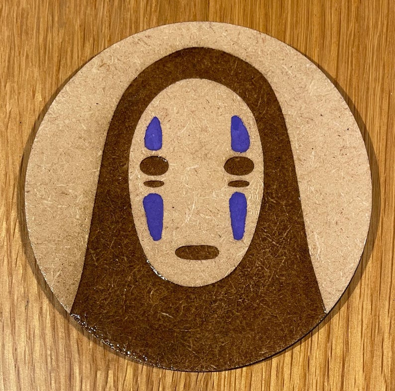 May include: A round, brown wooden coaster featuring a brown and purple No Face character from the film Spirited Away.