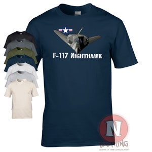 以下が含まれることがあります： F-117 Nighthawkステルス戦闘機が白で描かれたネイビーブルーのTシャツ。機体は正面を向いており、尾部にアメリカ空軍の星が描かれている。
