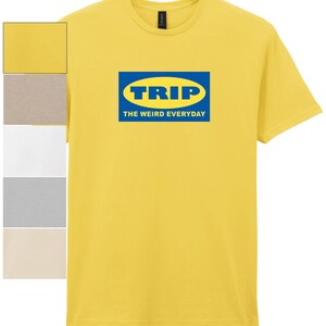 Puede incluir: Camiseta amarilla con un logotipo azul y amarillo que dice "TRIP" en un óvalo azul, con el texto "THE WEIRD EVERYDAY" debajo. La camiseta tiene mangas cortas y cuello redondo.