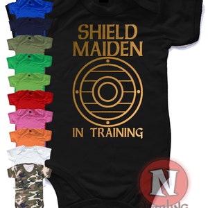 Op de afbeelding: Zwarte baby romper met gouden tekst die "Shield Maiden In Training" zegt en een gouden schildontwerp. De romper wordt in verschillende kleuren getoond.