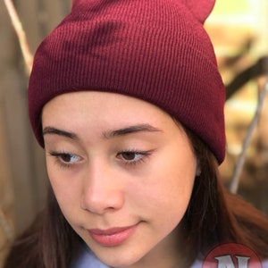 May include: A close-up of a person wearing a maroon beanie. The beanie is made of a soft, knitted fabric. The person is looking down and has a neutral expression.