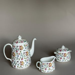 May include: A white porcelain tea set with a floral pattern. The set includes a teapot, creamer, and sugar bowl. The teapot has a curved spout and handle, and the sugar bowl has a lid. The floral pattern features pink, yellow, and blue flowers.