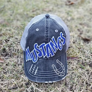 May include: A grey and white trucker hat with a blue embroidered "MUSTANGS" logo. The hat has a distressed look and a horseshoe design on the back.