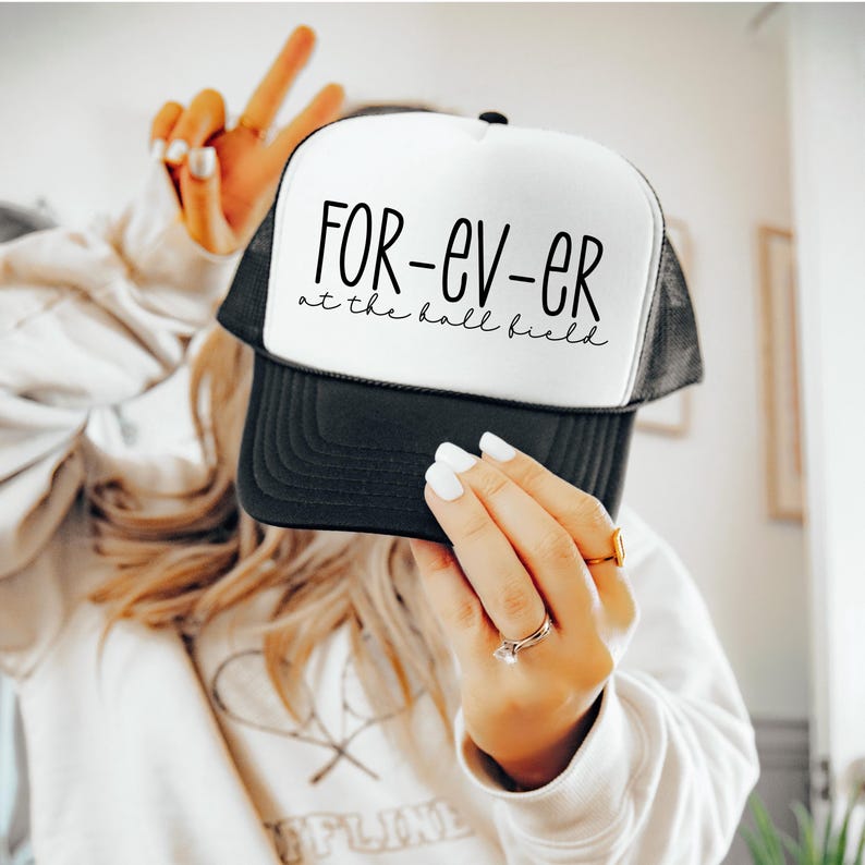 K&ouml;nnte beinhalten: Eine schwarz-wei&szlig;e Trucker-Kappe mit dem Text "FOR-EV-ER at the ball field" in schwarzer Schrift.