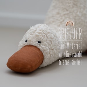 Könnte beinhalten: Ein weißer Plüsch-Enten mit einem braunen Schnabel und schwarzen Augen. Die Ente ist ein Musikspielzeug mit dem Text "SPIELUHR Bio-Material Melodieauswahl personalisierbar" an der Seite.