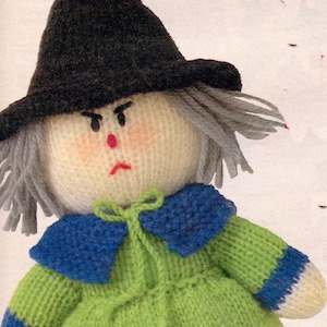 May include: A knitted witch doll with a black hat, gray hair, a green dress with blue stripes, and a blue collar. The doll has a painted face with a red nose and a frown.