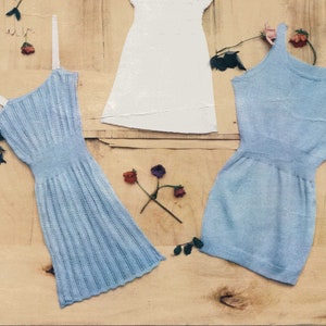Puede incluir: Dos vestidos de bebé de punto, uno blanco y otro azul claro, expuestos sobre un fondo de madera con flores rosas y rojas. Los vestidos son sin mangas y tienen un diseño sencillo. El texto "LAVENDA HAND KNIT 901 SIXPENCE" es visible en la parte superior de la imagen.