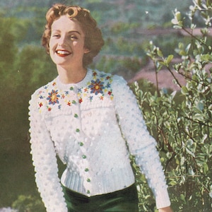 May include: A white cardigan with a floral design on the front. The cardigan is made with a textured stitch and has green buttons. The model is wearing a green skirt.
