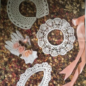May include: A collection of white crocheted collars and a glove adorned with pink roses. The collars are in various shapes, including a rounded collar, a circular collar, and an oval collar. The background features dried flowers and a pink ribbon.