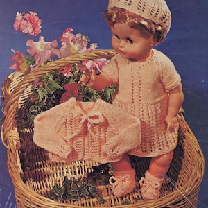 May include: A pink knitted doll dress and cardigan set with a matching pink knitted hat. The doll is wearing the outfit and is sitting in a wicker basket with pink flowers.