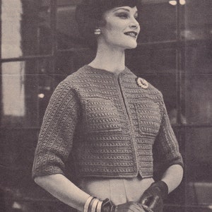 May include: A close-up of a woman wearing a gray crocheted cardigan with a button closure. She is wearing a skirt and gloves. The cardigan has a textured pattern and is cropped at the waist.