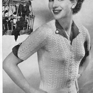 May include: A woman wearing a white knitted cardigan with a black collar and buttons. The cardigan has a chevron pattern and short sleeves. The woman is standing in front of a wicker basket.