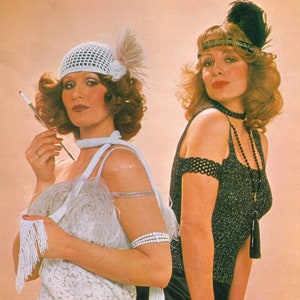 Può includere: Due donne vestite in stile flapper degli anni '20. Una donna indossa un cappello bianco all'uncinetto e un vestito bianco con un boa di piume. L'altra donna indossa un top nero con paillettes, una collana di perle nere e un cerchietto con piume nere.