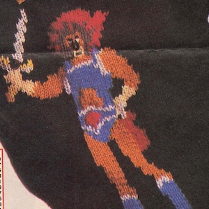 Puede incluir: Un suéter de punto negro con una imagen de punto de colores de una figura similar a un león que sostiene una espada. La figura lleva un atuendo azul y blanco con un corazón rojo en el pecho.