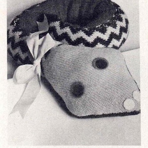 May include: A black and white knitted hat with a zig-zag pattern and a white ribbon tied around it. The hat is sitting on top of a grey knitted pillow with black buttons.