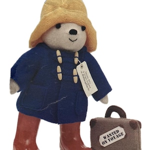 May include: A knitted Paddington Bear toy, wearing a yellow hat, blue coat, and red wellington boots. The bear has a small suitcase with a "WANTED ON VOYAGE" label. A white tag hangs from the coat.
