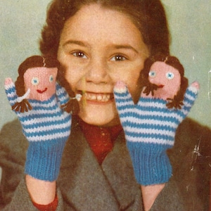 May include: A young girl wearing a grey coat is smiling and holding two knitted finger puppets. The puppets are pink and blue with white stripes and have brown hair and blue eyes.